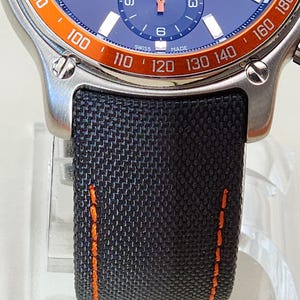 Op de afbeelding: Close-up van een roestvrijstalen polshorloge met een blauwe wijzerplaat en oranje lunette. Het horloge heeft een zwarte stoffen band met oranje stiksels. De wijzerplaat toont de dag en tijd.