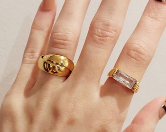 May include: A hand with long, almond-shaped nails with a nude base and black abstract designs. The hand is wearing a gold chain bracelet and two gold rings, one with a clear stone.