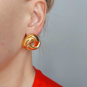 Peut inclure: Boucles d'oreilles en forme de nœud dorées. Les boucles d'oreilles sont grandes et ont un design torsadé.
