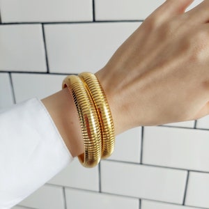 Może przedstawiać: Dwa złote bransoletki w kształcie węża, ułożone na nadgarstku osoby. Bransoletki są wykonane z elastycznego metalu i mają błyszczące wykończenie.