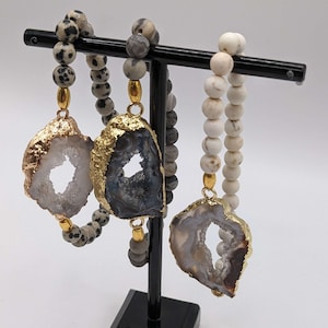 Peut inclure: Trois colliers de perles avec des pendentifs en agate à bordure dorée sont exposés sur un présentoir noir. Les perles sont dans des tons de gris, de blanc et de noir moucheté. Les pendentifs en agate ont un centre naturel rempli de cristaux.