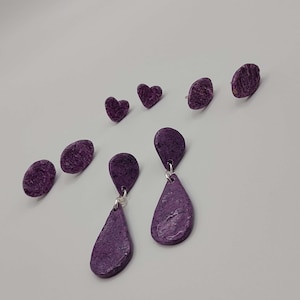 May include: A collection of purple earrings in various shapes. The set includes two pairs of stud earrings, two heart-shaped studs, and a pair of teardrop dangle earrings. The earrings have a textured surface and are displayed on a white background.