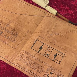 May include: A brown parchment paper featuring a hand-drawn architectural plan of a building and grounds, labeled A Plan of part of Mount Vernon Lands, with a white and brown tobacco pipe resting on the top edge of the document.