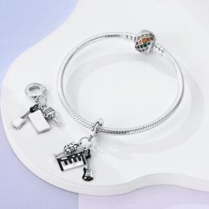 May include: A silver charm bracelet with a colorful charm and two music-themed charms. One charm features a microphone, a keyboard, and a guitar. The other charm features a guitar, a microphone, and a blank space.