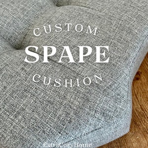 May include: A gray, tufted cushion with the words "CUSTOM SPACE CUSHION" in white lettering. The cushion has a textured fabric and is likely for a chair or seating area. The background is a wooden floor.