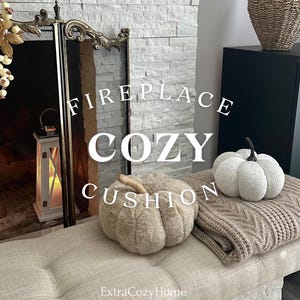 May include: A fireplace with a cozy cushion in the shape of a pumpkin. The cushion is brown and fluffy. There is a white pumpkin on the right side of the cushion. The text "Fireplace Cozy Cushion" is written on the image. The text "ExtraCozyHome" is written at the bottom of the image.