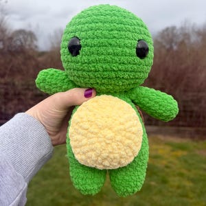 Pode incluir: Um peluche de tartaruga verde de crochê feito à mão, com barriga amarela e olhos pretos. A tartaruga tem braços e pernas pequenos e é feita de um fio macio e texturizado. O peluche é segurado contra um fundo desfocado ao ar livre.