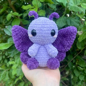 May include: A handmade, plush butterfly in shades of purple. The butterfly has large, dark eyes, small antennae, and detailed wings. The soft, textured material gives it a cuddly appearance. The butterfly is a charming, whimsical creation.