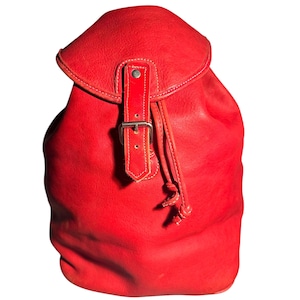 May include: A bright red leather backpack featuring a fold-over flap with a single vertical strap, a metal buckle closure, and a drawstring cord. The bag has a smooth, matte texture and a rounded silhouette with visible stitching details.