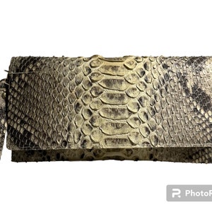 Könnte beinhalten: Eine beige Clutch aus Schlangenleder mit einem langen Riemen. Die Clutch hat einen Klappverschluss und ein strukturiertes Schlangenledermuster.