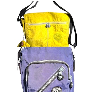 Op de afbeelding: Twee crossbody tassen, één gele en één paarse, met zwarte bandjes. De gele tas heeft een voorvak en een apen sleutelhanger. Beide tassen hebben een ritssluiting en een rond logo.