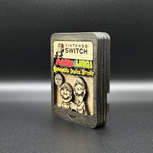 Könnte beinhalten: Ein hölzerner Nintendo Switch-Spielekartuschen-Kasten mit den Figuren Mario und Luigi aus dem Spiel "Mario & Luigi: Bowser's Inside Story". Der Kasten ist dunkelbraun mit einem hellbraunen Innenraum und einem schwarzen Nintendo Switch-Logo.