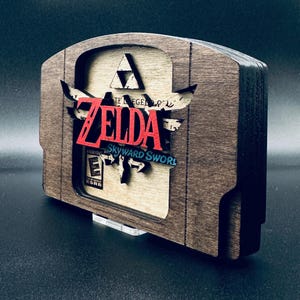 May include: Wooden game cartridge display featuring the Zelda: Skyward Sword logo in red and blue. The display has a dark brown frame with a light brown interior. The ESRB rating is visible. A unique piece for video game enthusiasts.