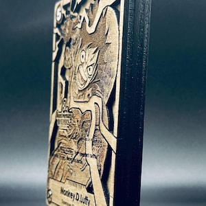 May include: A layered wooden art piece featuring a character from the One Piece anime series. The artwork is in shades of brown and black, with the character's image and text details carved into the wood. The piece is displayed on a clear stand.