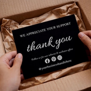 May include: A black card with white text that reads "WE APPRECIATE YOUR SUPPORT" and "thank you" in cursive. The card also includes social media icons and the handle @yourbusinesshandlehere. The card is held in a cardboard box.