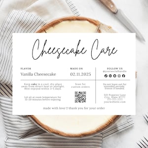 May include: A partially sliced vanilla cheesecake with a golden-brown crust, presented on a white surface with a striped cloth. A white card displays "Cheesecake Care" instructions, including flavor, date made, and care tips. A knife is visible.