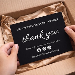 Puede incluir: Una tarjeta negra con texto blanco que dice "WE APPRECIATE YOUR SUPPORT" y "thank you" en cursiva. La tarjeta tambi&eacute;n incluye iconos de redes sociales y el identificador @yourbusinesshandlehere. La tarjeta se sostiene en una caja de cart&oacute;n.