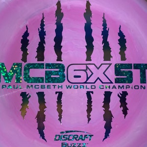 May include: A pink disc golf disc with a black and silver claw mark design. The disc has the text "MCB6XST PAUL MCBETH WORLD CHAMPION" in a glittery, iridescent font.