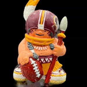 May include: A figurine of a Native American figure in a football helmet, holding a football and a spear. The figure is wearing a burgundy helmet with yellow stripes, a yellow scarf, and a red football. The figurine is on a black background.