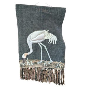 May include: A rectangular dark gray wall hanging featuring an embroidered white crane standing in a grassy area. The crane has a long neck and slender legs. The bottom edge of the textile is finished with a long, multi-colored fringe.