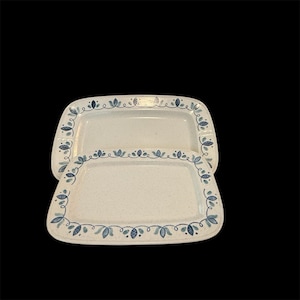 May include: Two rectangular serving platters with a speckled white surface. Each platter has a decorative border of blue leaves and dots. The platters are stacked on top of each other against a black background.