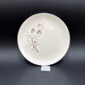 May include: A round, off-white ceramic plate featuring a delicate, dark brown botanical illustration of leaves and flowers arranged in a curved pattern on the left side, finished with a thin, metallic silver rim around the outer edge.