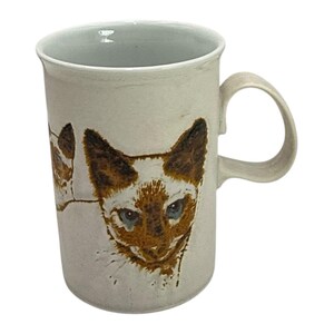 May include: Cream-colored ceramic mug featuring a Siamese cat design. The cat's face is brown and white with blue eyes. The mug has a curved handle and a simple, elegant design, perfect for coffee or tea.