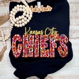 Puede incluir: Sudadera negra con las palabras "Kansas City CHIEFS" en oro y purpurina roja. Un collar de cuentas de madera y una etiqueta de madera que dice "Lala Lou's" están en la parte superior izquierda.