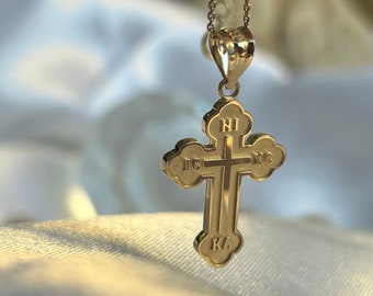 May include: A gold-colored pendant in the shape of a cross with Greek lettering. The cross is on a white background.