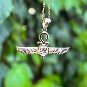 May include: Gold pendant necklace featuring an ankh symbol with wings and the Eye of Horus. The pendant is on a chain against a green leafy background.