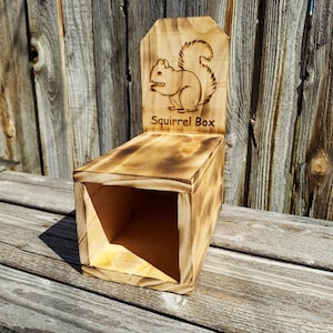 May include: A wooden squirrel box with a burnt finish. The box has a rectangular body and a backboard with a squirrel engraving and the words "Squirrel Box". The box is made of light-colored wood.