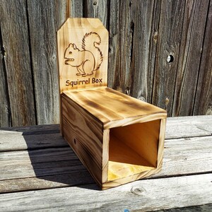 May include: A wooden squirrel box with a light brown finish. The box has a rectangular body and a backboard with a carved squirrel design and the words "Squirrel Box". The box is made of wood and has a rustic appearance.