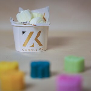 Könnte beinhalten: Eine weiße Tasse mit einem goldenen "ZK Candle Co." Logo, gefüllt mit kleinen weißen Wachs-Schmelzen. Die Tasse ist umgeben von bunten Wachs-Schmelzen in verschiedenen Formen.
