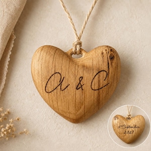 May include: A heart-shaped wooden ornament featuring a natural wood grain finish and a light brown twine hanger. The front is engraved with the letters a & C in a cursive script. A smaller inset shows the back engraved with 5th September 2021.