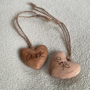 Op de afbeelding: Twee houten hartvormige ornamenten met touwhangers. Eén hart heeft de inscriptie "A&K", de andere "E&R". De ornamenten zijn lichtbruin van kleur en liggen op een getextureerd, lichtgrijs oppervlak.
