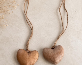 May include: Two heart-shaped ornaments carved from light brown wood, each featuring a smooth, polished surface with visible natural grain patterns. Each heart is attached to a thin, twisted jute twine loop for hanging, showcasing a simple design.