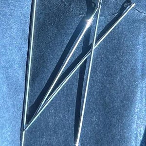 Puede incluir: Primer plano de varias agujas de coser plateadas sobre una superficie azul oscuro. Las agujas son finas y tienen un peque&ntilde;o ojo. Est&aacute;n dispuestas de forma ligeramente superpuesta, proyectando sombras.