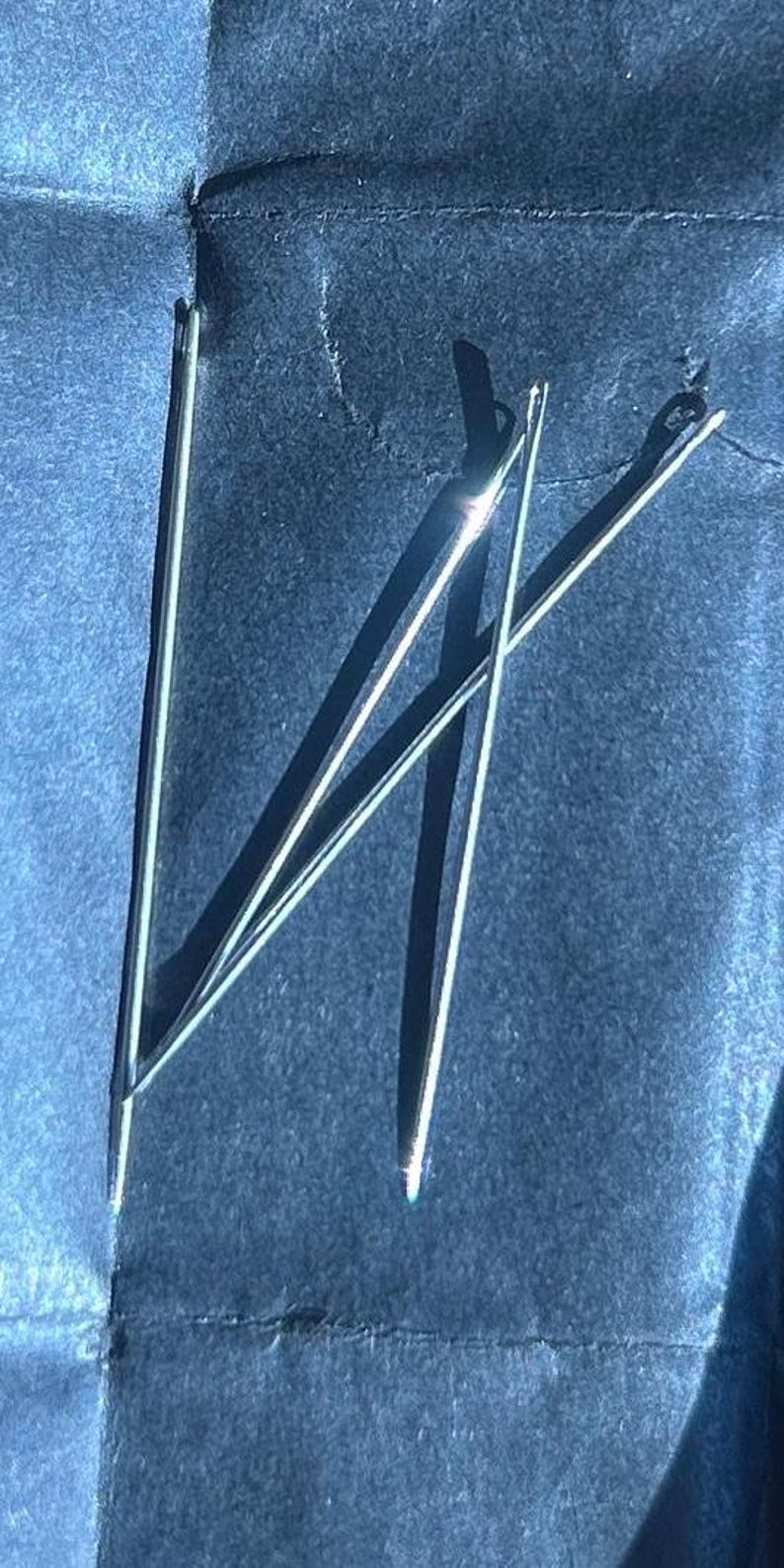 Puede incluir: Primer plano de varias agujas de coser plateadas sobre una superficie azul oscuro. Las agujas son finas y tienen un peque&ntilde;o ojo. Est&aacute;n dispuestas de forma ligeramente superpuesta, proyectando sombras.
