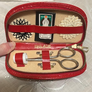 Puede incluir: Estuche de costura rojo de polipiel abierto con cremallera dorada. En el interior, accesorios de costura, incluyendo unas tijeras peque&ntilde;as, agujas y carretes de hilo decorativos. El kit est&aacute; marcado "GROUND LEATHER CASE MADE IN AUSTRIA."