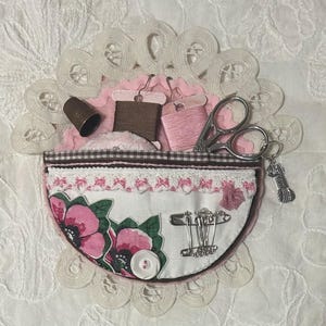 May include: A handmade sewing kit with a floral print pocket, lace trim, and a brown gingham border. The kit contains spools of thread, scissors, safety pins, a thimble, and a charm. The background is a white, textured fabric.
