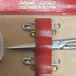 Puede incluir: Primer plano de un estuche de cuero rojo con el texto "GROUND LEATHER CASE MADE IN AUSTRIA". El estuche contiene unas tijeras plateadas, dos imperdibles y una correa de cuero roja.