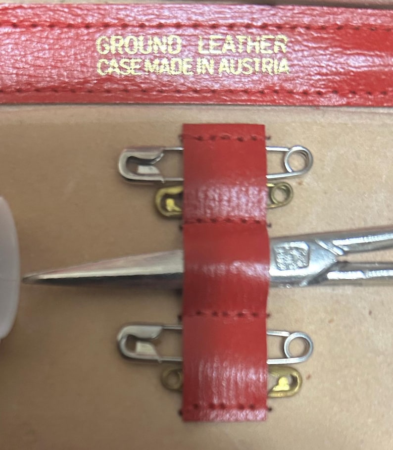 Puede incluir: Primer plano de un estuche de cuero rojo con el texto "GROUND LEATHER CASE MADE IN AUSTRIA". El estuche contiene unas tijeras plateadas, dos imperdibles y una correa de cuero roja.