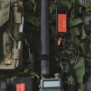 May include: A black two-way radio with a keypad and a small screen. The radio is attached to a black strap and is sitting on a green camouflage fabric. There is a small black rectangle with red text that says "SIGNAL MIRROR" and a black rectangle with red text that says "KIT FIELD ONE" in the image.