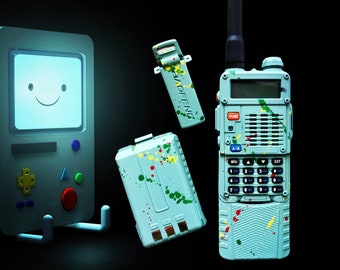 Puede incluir: Una radio de dos vías de color azul verdoso y blanco con una antena negra, una batería azul verdoso y blanca y un clip azul verdoso y blanco. La radio y los accesorios están cubiertos de salpicaduras de pintura de colores. El texto "FENG BROS" está impreso en amarillo sobre un fondo negro.