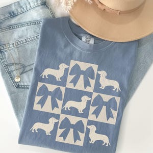 May include: A light blue t-shirt with a white graphic of a checkered pattern featuring dachshunds and bows. The shirt has the text "COMFORT COLORS" on the neck.
