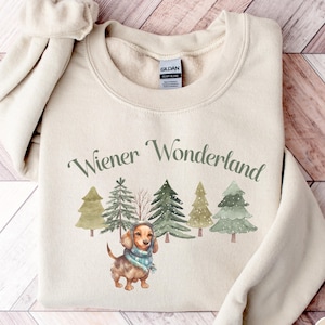May include: A sand-colored sweatshirt with the text "Wiener Wonderland" in green script. Below the text is a watercolor illustration of a dachshund wearing a hat and scarf, with Christmas trees in the background.