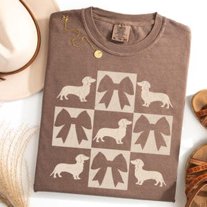 May include: A brown t-shirt with a pattern of white dachshunds and bows. The shirt has a tag that says "Comfort Colors" and "XL - TG - EG".