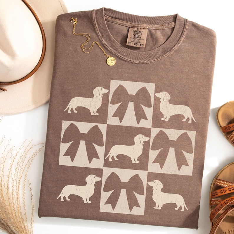 May include: A brown t-shirt with a pattern of white dachshunds and bows. The shirt has a tag that says "Comfort Colors" and "XL - TG - EG".