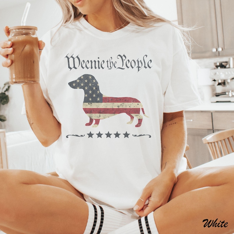 May include: White t-shirt with the text "Weenie the People" in a vintage font. The shirt features a dachshund dog design with an American flag pattern. Below the dog are five stars and a decorative line.