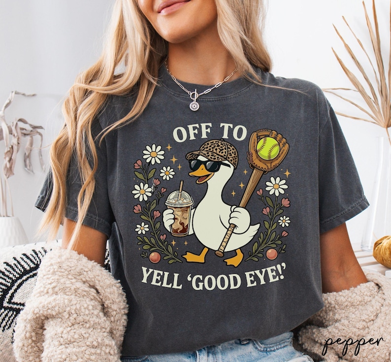 May include: A charcoal grey t-shirt featuring a graphic of a white duck wearing a leopard print baseball cap and sunglasses. The duck holds an iced coffee and a softball bat with a ball. Text reads: OFF TO YELL 'GOOD EYE!' surrounded by flowers.